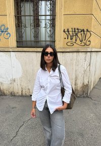 Vrouw met een zonnebril, witte shirt en grijze spijkerbroek die op een stoep in de stad staat nabij een geel gebouw met graffiti en vergrendelde ramen.