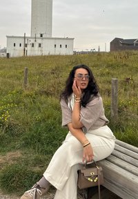 Svetlo sivi široki hlače, taupe oversized pulover, smeškan torba z zlatim okovjem in rjavi športni čevlji. Zeleno travo in oblačno ozadje.