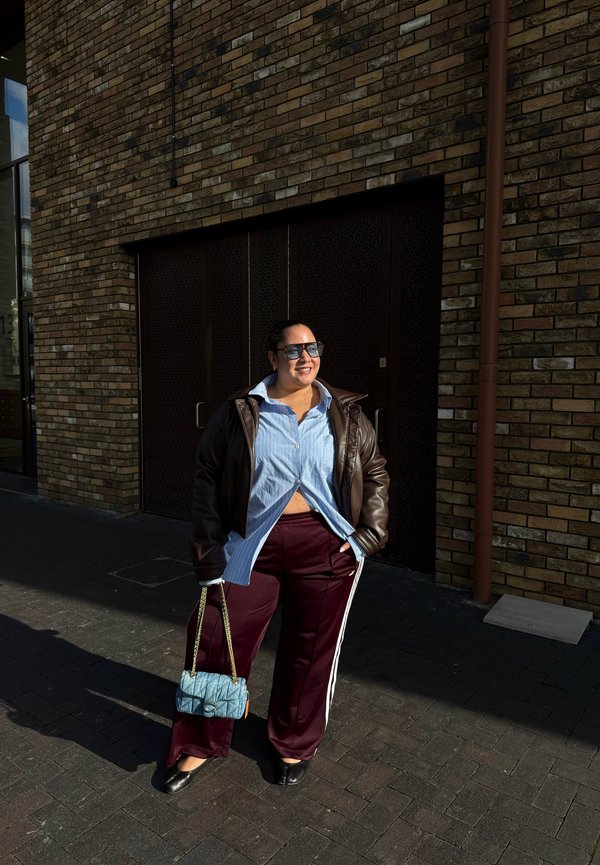 Bruine leren jas, lichtblauw gestreept overhemd, kastanjebruine broek met witte strepen, zwarte flats en een blauwe gewatteerde handtas. Staand buiten.