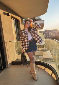 Černobílá kostkovaná bunda, bílé tričko, džínová sukně, béžové kovbojské boty, držící černou kabelku, stojící na balkóně na slunečním světle.