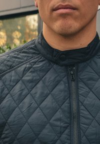 Μαύρο επενδυμένο μπουφάν με ψηλό γιακά, κλείσιμο με κουμπιά και μπροστινό φερμουάρ. Λείο ύφασμα με μοτίβο σε σχήμα διαμαντιού.