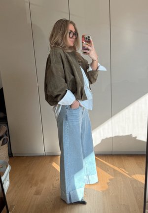 Chaqueta oversized de ante marrón sobre una camisa blanca, combinada con jeans de pierna ancha azul claro. Presenta una textura suave y puños enrollados.