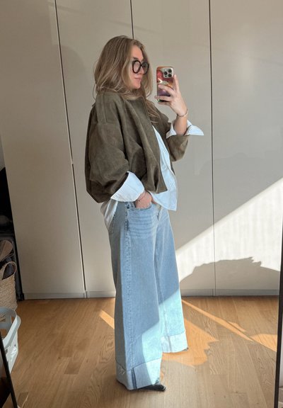 Chaqueta oversized de ante marrón sobre una camisa blanca, combinada con jeans de pierna ancha azul claro. Presenta una textura suave y puños enrollados.
