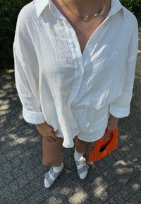 Λευκό, υφασμάτινο πουκάμισο με κουμπιά, συνδυασμένο με αντίστοιχο σορτς. Πλεκτή τσάντα σε πορτοκαλί απόχρωση και γκρι σανδάλια με λουριά.