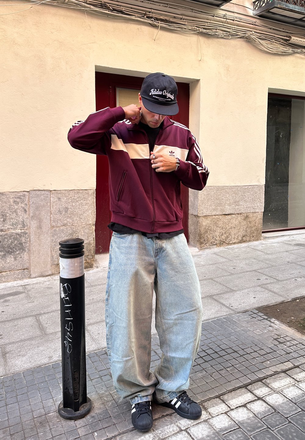 Jachetă crem cu fermoar de culoare maron, blugi mari albaștri și adidași negri cu dungi albe, stând lângă un stâlp metalic negru.