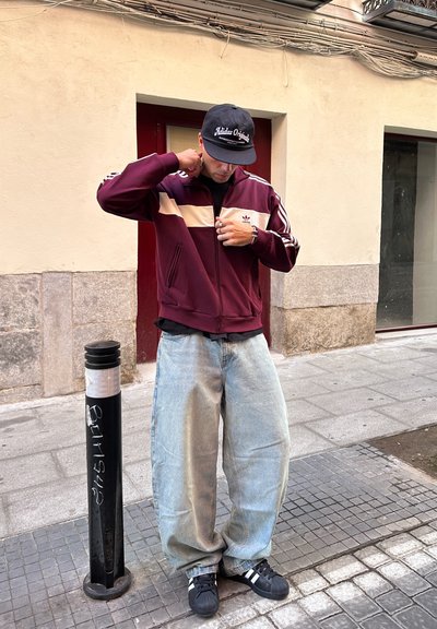 Chaqueta de tiro marón con raya crema, jeans oversize azules y zapatillas negras con rayas blancas, de pie cerca de un poste de metal negro.