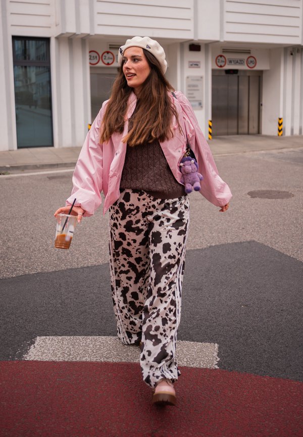 Joven mujer camina por la calle con una chaqueta rosa, boina blanca, pantalones negros con estampado, sosteniendo una bebida fría y un bolso con un llavero de peluche morado.