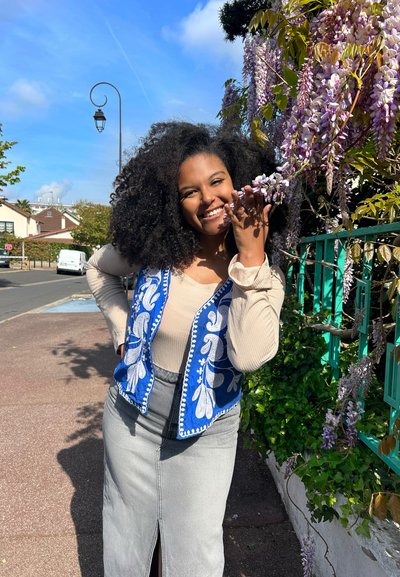 Chaleco azul bordado con patrones florales blancos sobre una blusa beige, acompañado de una falda de mezclilla gris y rodeado de flores moradas y vegetación.