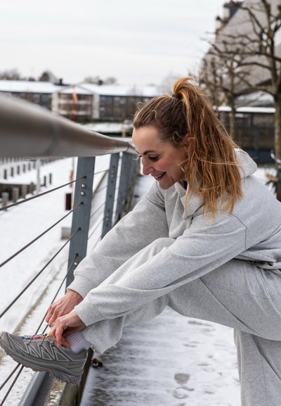 Sudadera gris claro y joggers, con un enfoque en zapatillas deportivas grises con acentos coloridos. El entorno presenta un paisaje exterior nevado.