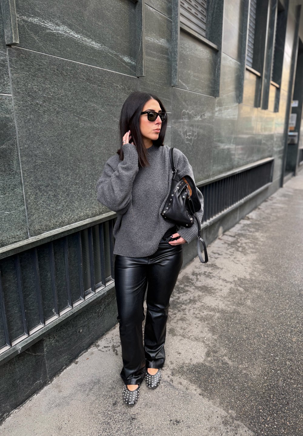 Szürke túlméretezett pulóver, fekete műbőr nadrág, fekete táska szegecsekkel, és ezüst részletekkel díszített hegyes orrú cipők egy városi utcán.