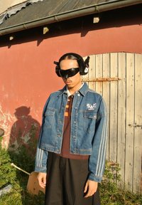 Τζάκετ από τζιν με μπλε απόχρωση, με δύο μπροστινές τσέπες, μεταλλικά κουμπιά και ριγέ λεπτομέρειες στα μανίκια. Φορεμένο πάνω από ένα καφέ πουκάμισο και μαύρα παντελόνια.