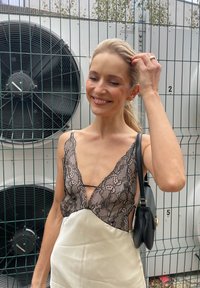 Zwarte kanten cami-top met diepe V-hals en satijnen ivoorkleurige rok. Model houdt een kleine zwarte handtas vast. Industrieel achtergrond met grote ventilatoren.