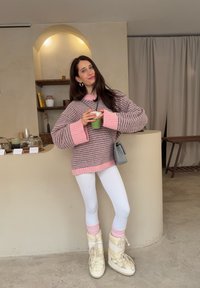 Jeune femme portant un pull à rayures roses, des leggings blancs et des bottes crème, tenant une boisson verte dans un café au décor minimaliste.
