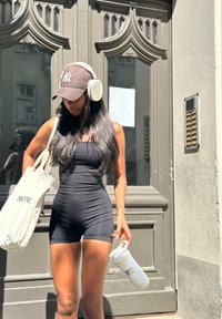 Μαύρο εφαρμοστό κορμάκι, μακριά μαύρα μαλλιά, λευκά ακουστικά, μαύρο καπέλο, κρέμ τσάντα με κείμενο και λευκό μπουκάλι νερού με λογότυπο, standing by a door.