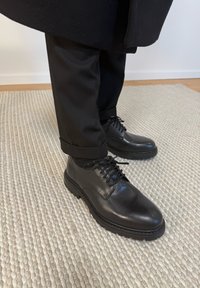 Μαύρα δερμάτινα παπούτσια με κορδόνια, φορεμένα με μαύρο παντελόνι και μαύρο παλτό πάνω σε υφασμένο μπεζ χαλί σε εσωτερικό χώρο.