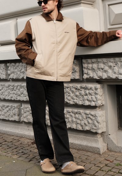 Chaqueta con cremallera en tonos beige y marrón, con cuello, combinada con jeans negros de pierna recta y zuecos de color marrón claro. Fondo de pared texturizada en gris.