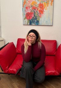 Rotes Kunstledersofa mit minimalistischem Design und einem schwarzen Metallrahmen. Getragen werden graue Nadelstreifenhose und ein bordeauxroter Rippstrickpullover.