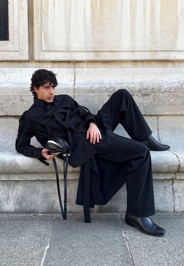 Hombre con atuendo negro reclinado en un banco de piedra, sosteniendo un bolso negro plisado con correas, pared de piedra texturizada al fondo.