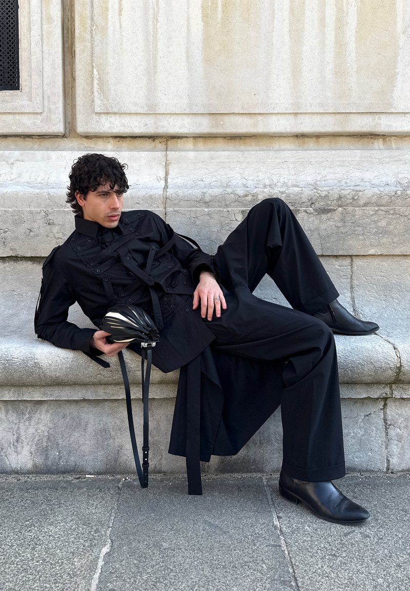 Man in black outfit reclining on stone bench, holding black pleated handbag with straps, textured stone wall in background.