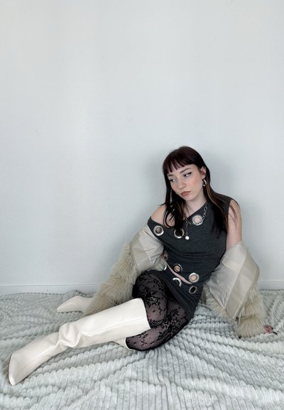 Vestido gris ajustado con cortes circulares, abrigo de piel sintética crema, medias de encaje negras y botas blancas hasta la rodilla sobre una manta gris texturizada.