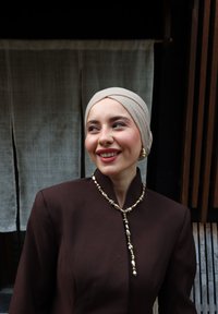 Smilende kvinde iført beige turban, mørkebrun jakke, guldperlesmykke og matchende øreringe, mod en stofbaggrund og træspær.