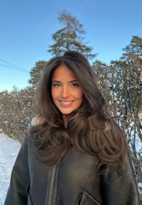 Femme aux longs cheveux ondulés souriante en manteau d'hiver, debout dans un paysage enneigé avec des buissons sans feuilles et des pins sous un ciel bleu clair.