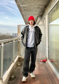 Casaco de couro preto, hoodie branco com logótipo, calças às riscas, gorro vermelho e sapatilhas brancas. De pé numa varanda com um fundo urbano visível.