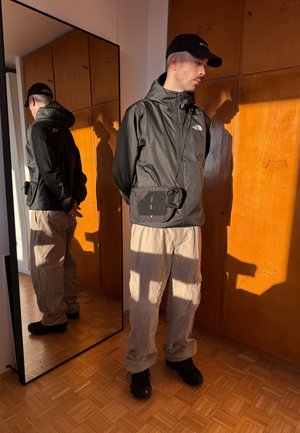 Hombre con chaqueta negra y gorra, pantalones cargo beige y bolso cruzado, de pie cerca de un armario de madera y un gran espejo bajo la cálida luz del sol.