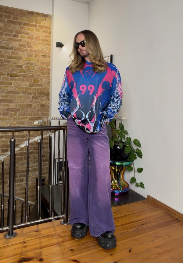 Una persona con el pelo largo y gafas de sol posa con una camiseta azul y rosa con el número 99, pantalones morados de pernera ancha y zapatos negros con plataforma.