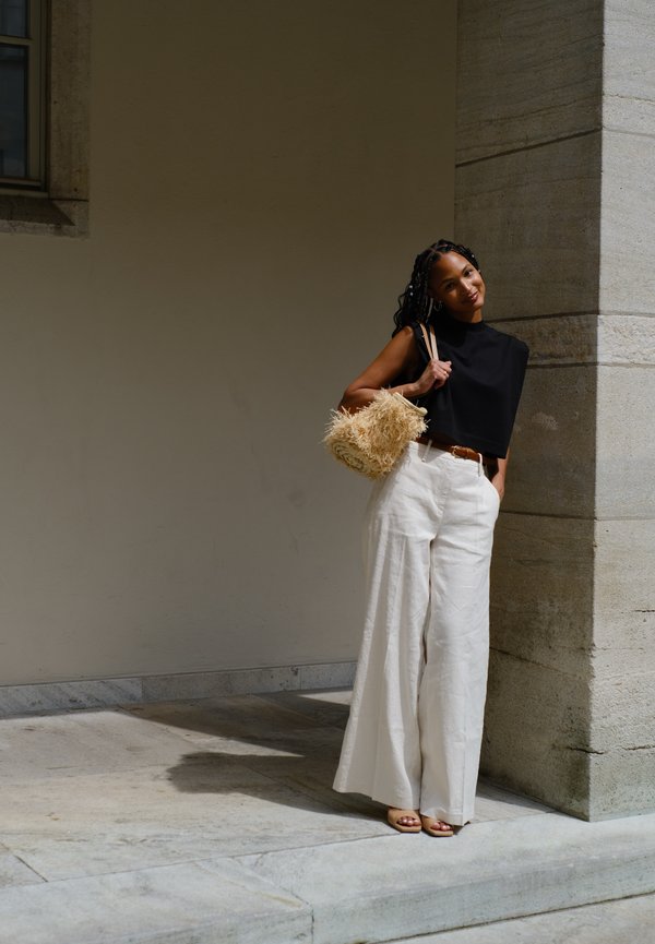 Zwarte cropped top, brede witte linnen broek en beige open teen sandalen. Houdt een gestructureerde beige strooien tas vast. Slanke silhouet, zachte texturen.