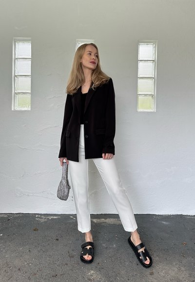 Blazer negro con dos bolsillos frontales, pantalones blancos y sandalias negras. Sostiene un pequeño bolso de mano con estampado. Fondo neutro con bloques de cristal.