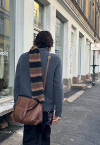Écharpe en laine à rayures marron, vertes et noires, portée sur un pull gris et associée à un sac messager en cuir marron. Cadre urbain.
