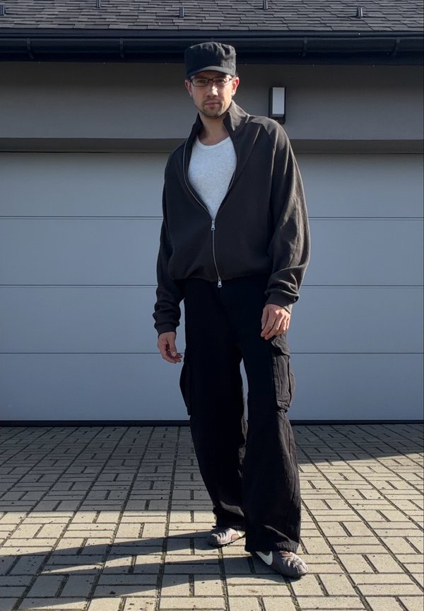 Hombre con gorra negra, gafas, chaqueta oscura con cremallera, camiseta blanca, pantalones cargo negros y zapatos Nike grises de pie en una entrada pavimentada.