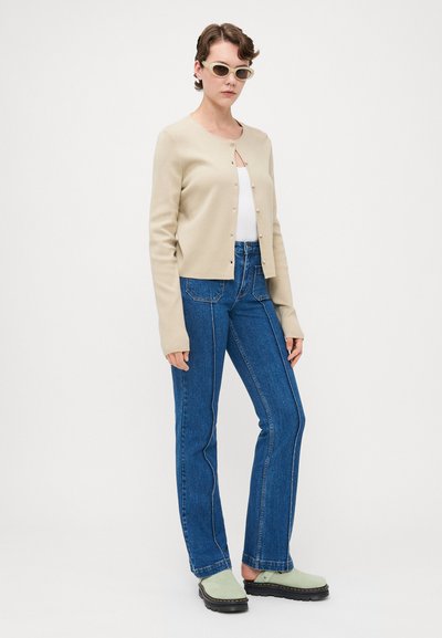 Cárdigan de punto beige con cuello redondo y botones de perlas, combinado con jeans acampanados azul. La modelo lleva zuecos verde claro y gafas de sol.