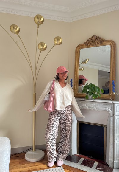 Mujer con gorra rosa, suéter blanco y pantalones de estampado de leopardo junto a una lámpara de pie dorada y un espejo ornamentado encima de una chimenea de mármol con planta.