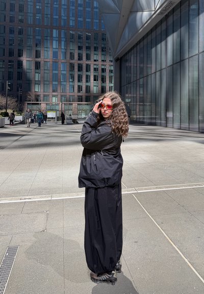 Mujer con cabello rizado que lleva una chaqueta negra, falda negra larga, patines en línea y gafas de sol rojas, está de pie en la acera de la ciudad cerca de un edificio moderno de vidrio.