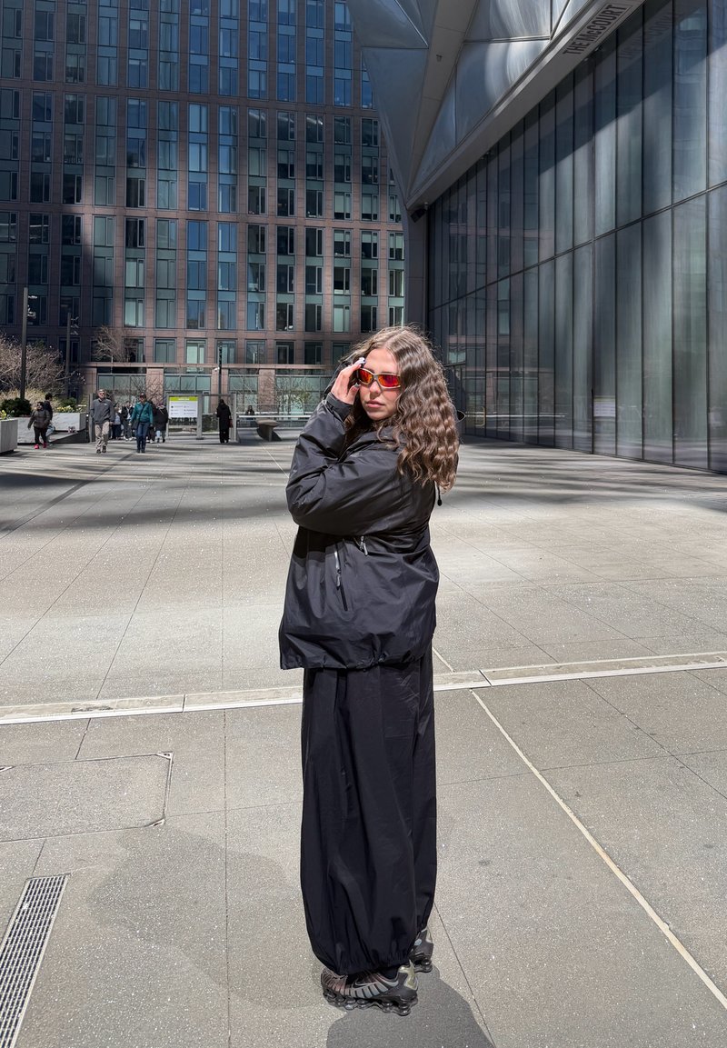 Femme aux cheveux bouclés portant une veste noire, une longue jupe noire, des rollers et des lunettes de soleil rouges, debout sur le trottoir de la ville près d'un bâtiment moderne en verre.