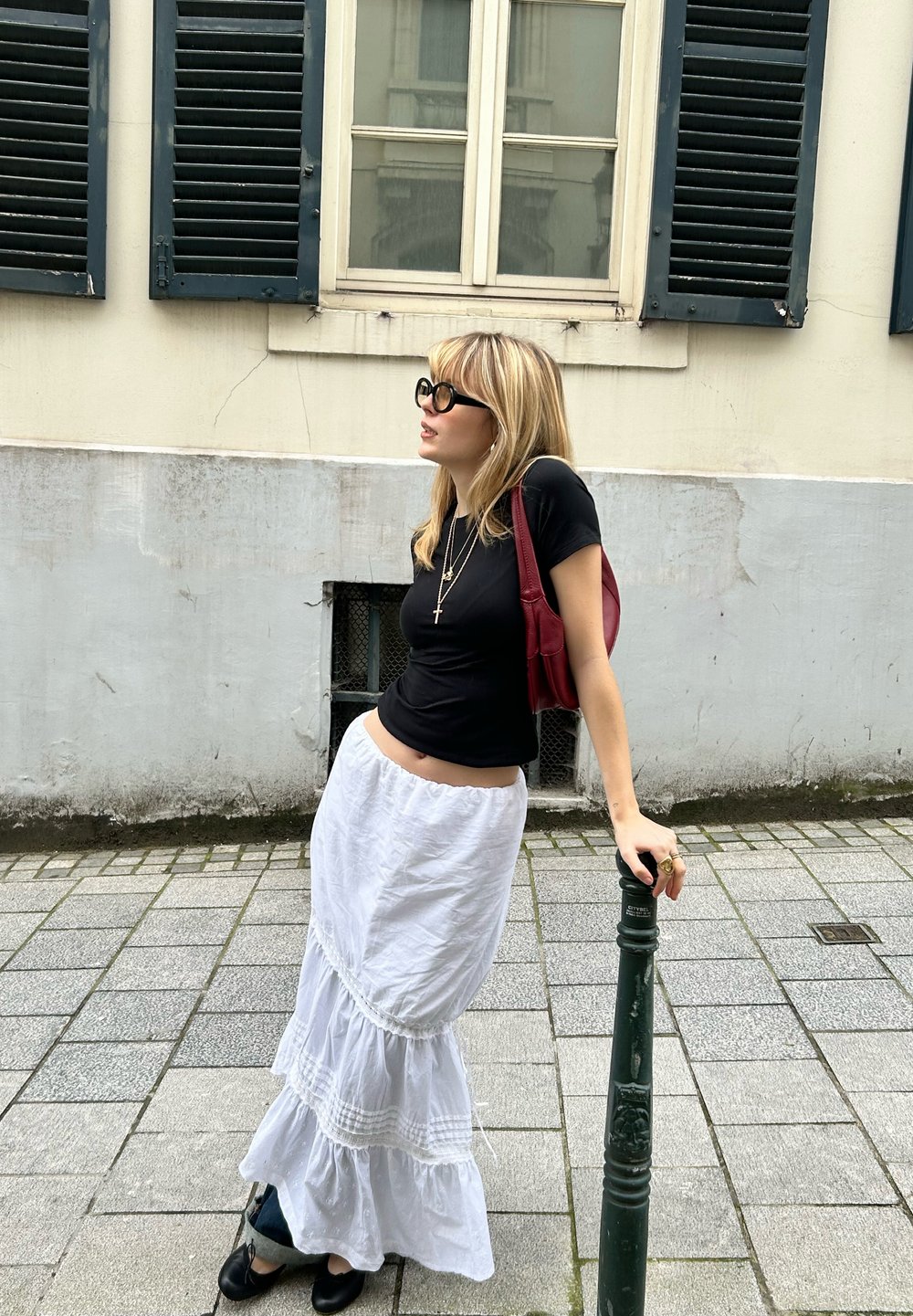 Μαύρη εφαρμοστή μπλούζα, λευκή φούστα με στρώσεις και δαντελωτές λεπτομέρειες, κόκκινη τσάντα ώμου και υπερμε�γέθη μαύρα γυαλιά ηλίου. Στέκεται δίπλα σε έναν πράσινο στύλο.