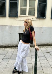 Μαύρη εφαρμοστή μπλούζα, λευκή φούστα με στρώσεις και δαντελωτές λεπτομέρειες, κόκκινη τσάντα ώμου και υπερμεγέθη μαύρα γυαλιά ηλίου. Στέκεται δίπλα σε έναν πράσινο στύλο.