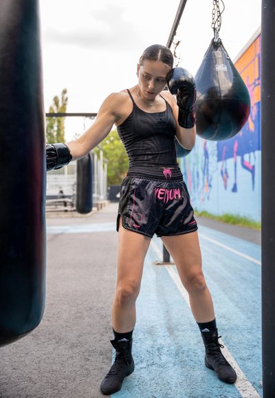 Camiseta sin mangas negra con un patrón sutil, pantalones cortos de boxeo negros con acentos rosas y logotipo, guantes de boxeo negros y zapatillas negras con cordones.