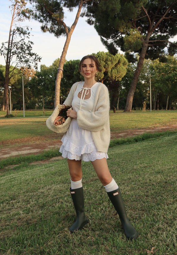 Vrouw in witte jurk en beige cardigan die een gevlochten tas met bloemen vasthoudt, staand op gras in een park met hoge bomen.
