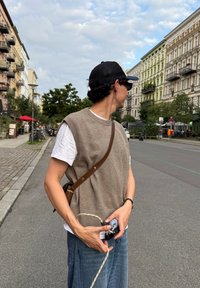 Een man met een pet, zonnebril en een vest houdt een oprolbare hondenlijn vast op een stadsstraat omringd door gebouwen en bomen.