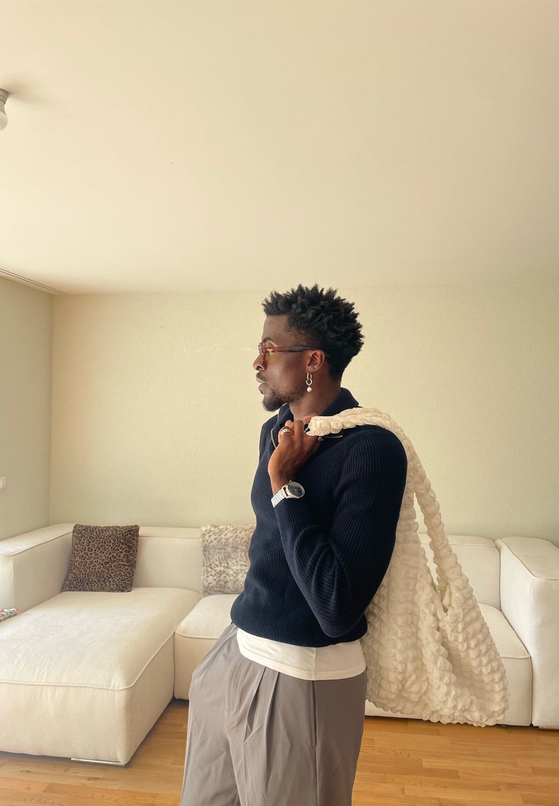 Man wearing glasses and watch stands in living room holding large textured white bag over shoulder.