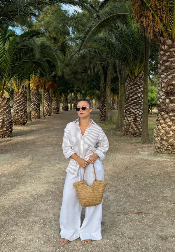 Femme portant des lunettes de soleil, une chemise rayée et un pantalon blanc, tenant un sac tressé, debout sur un chemin bordé de palmiers dans un cadre extérieur ensoleillé.