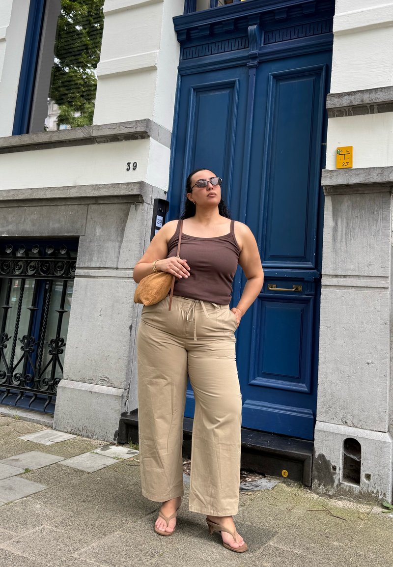 Femme portant des lunettes de soleil, un débardeur marron, un pantalon beige et des sandales, debout avec une main dans la poche et la sangle du sac sur l'épaule près d'une porte bleue.