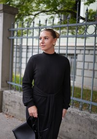 Svart ribt kjole med lange ermer, høy hals og knyttet livdetalj. Avsluttet med en elegant svart veske og gulløredobber.