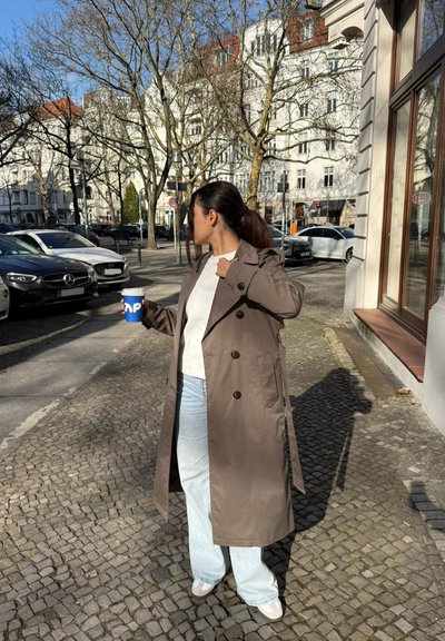 Un largo abrigo beige con botones oscuros, llevado sobre una blusa blanca y jeans de un azul claro. Sosteniendo una taza de café azul, de pie en una calle de adoquines.
