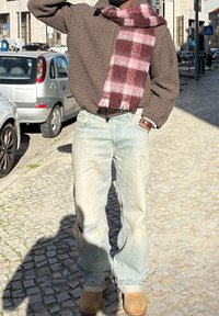 Chemise surdimensionnée à carreaux marron, écharpe à carreaux bordeaux et rose, jean délavé bleu clair, bottes beige, debout sur une rue pavée.