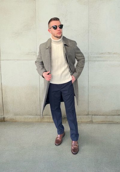 Abrigo de espiga gris, suéter de cuello alto crema, pantalones azul marino y mocasines de cuero marrón. Las gafas de sol completan el conjunto. Texturas suaves.