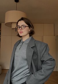 Blazer gris oversize porté sur un polo gris. Les lunettes sont foncées, et le d écor comporte un grand abat-jour au-dessus. Murs marron et sol clair.